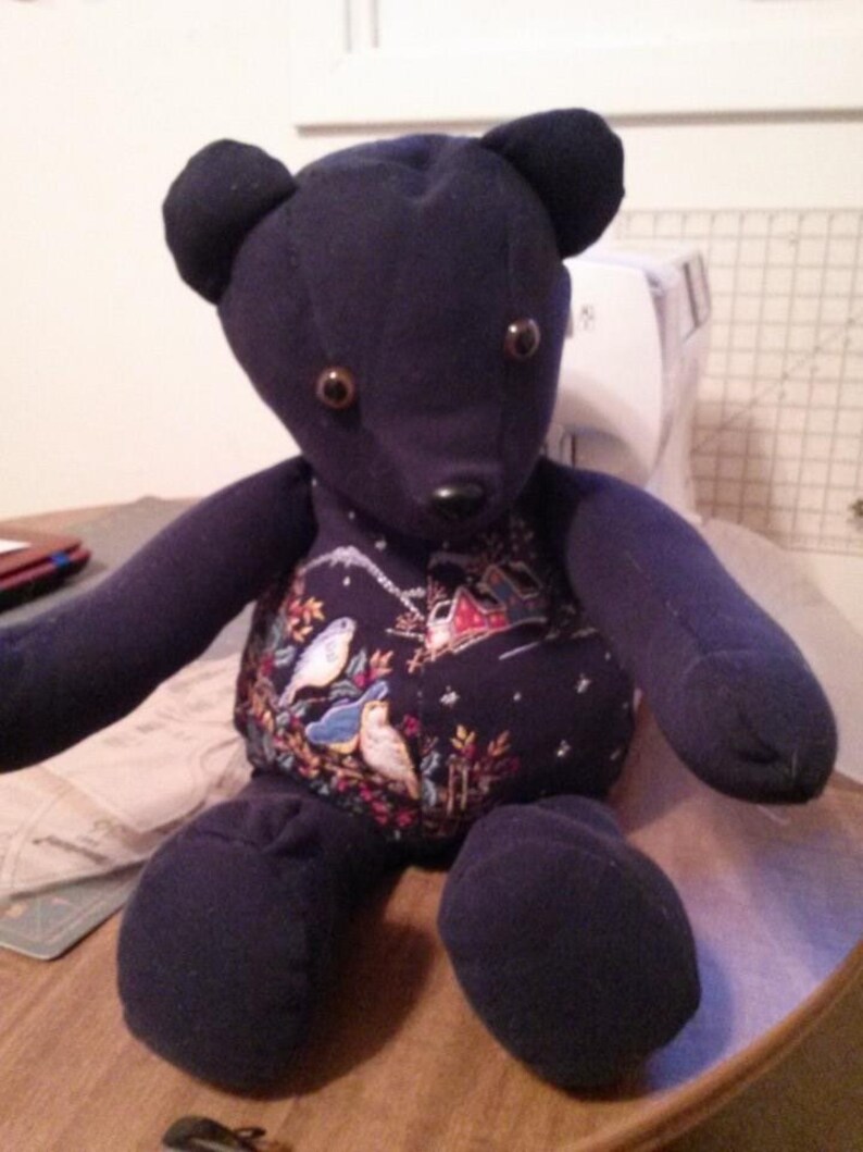 May include: A dark blue teddy bear with brown button eyes and a winter-themed fabric pattern on its chest. The bear is sitting on a wooden surface.