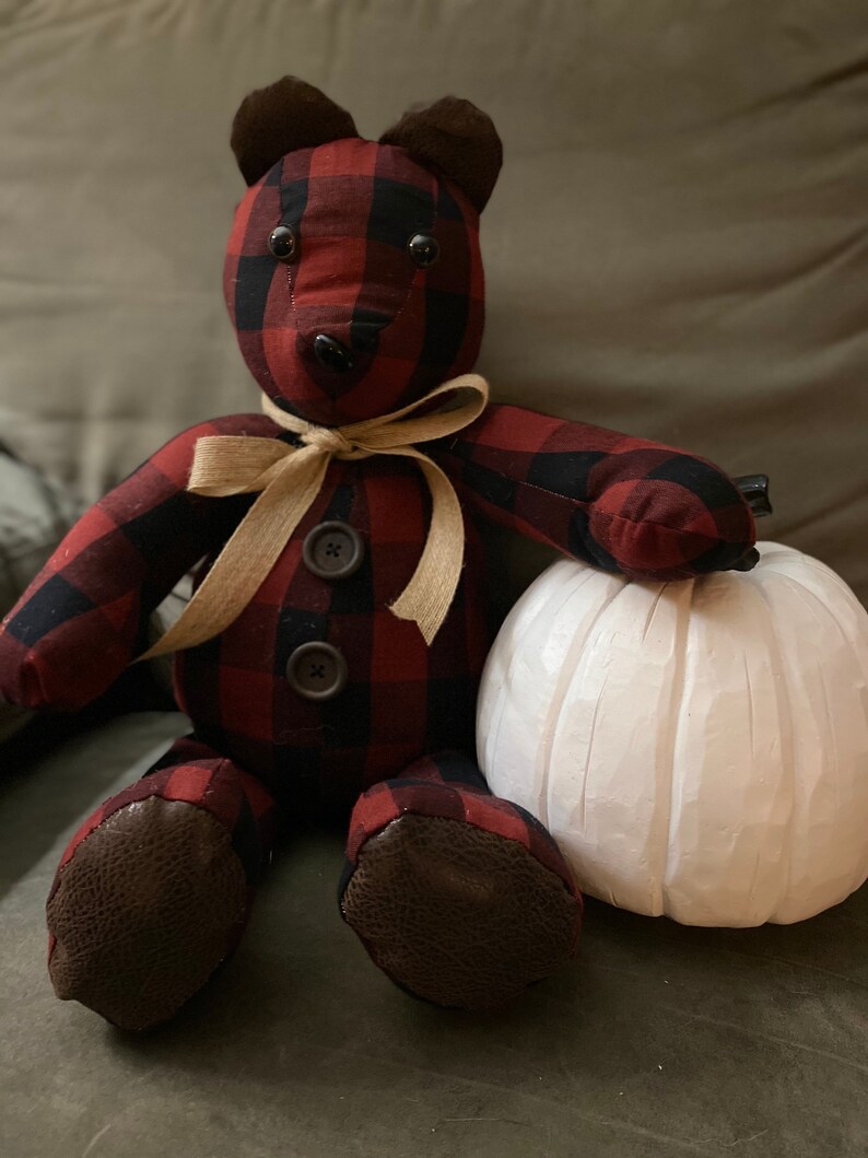 May include: A red and black plaid teddy bear with brown ears and black button eyes. The bear is wearing a tan ribbon around its neck and is sitting next to a white pumpkin.