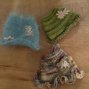 Puede incluir: Tres prendas de punto para muñecas en diferentes colores y estilos. Un gorro azul claro peludo, un gorro verde con un adorno floral y un vestido a rayas multicolor con un dobladillo con volantes.