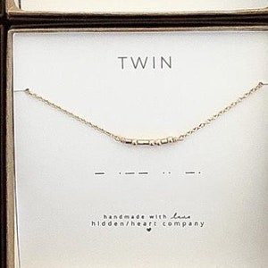 May include: Two gold necklaces in boxes with the word "TWIN" on the front. The necklaces are made of small gold beads and have a simple design. The boxes are brown with a gold trim.