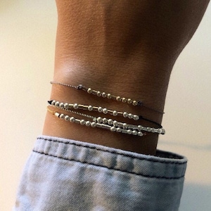 Peut inclure: Quatre bracelets délicats avec de petites perles dorées et argentées sont empilés sur un poignet. Les bracelets sont enfilés sur de fins cordons de différentes couleurs, dont le noir et le marron. Le poignet est couvert d'une chemise en jean bleu clair.