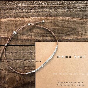 Peut inclure: Un bracelet en cordon marron avec des perles argentées et un charme en perle argentée. Le bracelet est sur une surface en bois brun avec une carte qui dit "mama bear" et "handmade with love hidden/heart company".