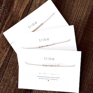 Peut inclure: Trois cartes blanches avec le mot "tribe" imprimé en noir sur chaque carte. Chaque carte a une fine chaîne dorée avec de petites perles dorées qui épellent le mot "tribe" en code Morse. La carte du bas a le texte "bijoux en code morse faits à la main hidden/heart company" imprimé en noir.