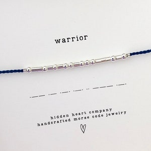 May include: A blue string bracelet with silver beads that spell out the word "warrior" in Morse code. The bracelet is packaged in a white box with the words "hidden heart company handcrafted morse code jewelry" printed on it.