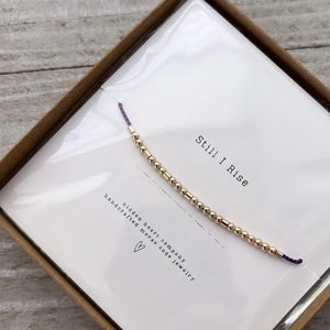 May include: A gold beaded bracelet with a purple string and a small card that says "Still I Rise" and "Hidden Heart Company handcrafted morse code jewelry".