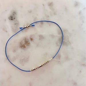 Peut inclure: Un bracelet en cordon bleu avec des perles dorées. Le bracelet est noué et a une petite perle dorée à l'extrémité.