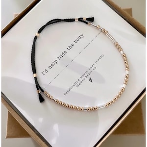 May include: A black string bracelet with gold and silver beads. The bracelet comes in a box with a card that reads "I'd help hide the body" and "handcrafted morse code jewelry hidden heart co."