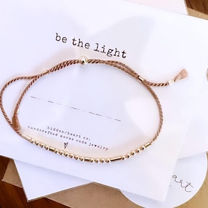 May include: A brown cord bracelet with a gold bead charm and a small silver bead. The bracelet has a message in gold beads that reads "be the light". The bracelet is on a white card with the text "hidden/heart co. handcrafted morse code jewellery".