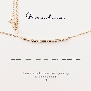 May include: Gold necklace with a delicate chain and a bar of small beads spelling out "Grandma" in Morse code. The necklace has a small heart charm. The text "handcrafted Morse code jewelry hidden/heart" is printed below the necklace.