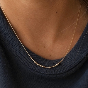May include: A gold chain necklace featuring a central segment composed of small round beads and short cylindrical bars arranged in a linear pattern. The delicate metal chain rests against the neck, showcasing a polished finish and a refined design.
