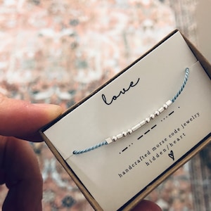 May include: A blue string bracelet with silver beads spelling out the word "love" in Morse code. The bracelet is in a box with the words "handcrafted morse code jewelry hidden/heart" printed on it.