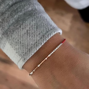 May include: A delicate bracelet featuring a thin gold bar and silver beads strung on a red cord. The bracelet is worn on a wrist, with a gray sleeve in the background. The bracelet is a minimalist piece of jewelry.