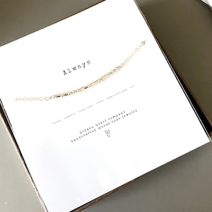Peut inclure: Une boîte blanche contenant un collier en chaîne dorée. Le collier est composé de petites perles dorées et épelle le mot "Always" en code Morse. La boîte porte l'inscription "hidden heart company handcrafted morse code jewelry".