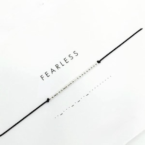 Peut inclure: Un bracelet en fil noir avec des perles argentées. Le mot "FEARLESS" est imprimé en lettres majuscules noires sur un fond blanc. Le bracelet est centré dans l'image.