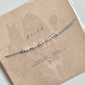 Peut inclure: Un bracelet en cordon gris avec des perles dorées qui épellent le mot "faith" en code morse. Le bracelet est emballé sur une carte brune avec le texte "hidden heart company handcrafted morse code jewelry" et un petit cœur.