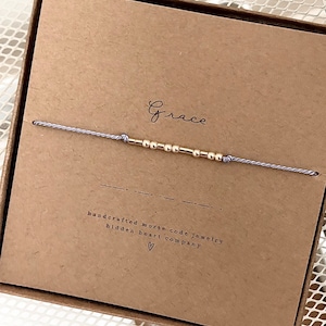 Peut inclure: Un bracelet en cordon gris avec des perles dorées qui épellent le mot "Grace" en code Morse. Le bracelet est présenté dans une boîte brune avec le texte "bijoux en code morse faits à la main hidden heart company" imprimé dessus.