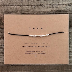 Peut inclure: Un bracelet en cordon noir avec des perles argentées qui épellent le mot "love" en code morse international. Le bracelet est fait à la main avec amour par hidden/heart company.