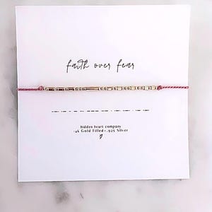 May include: A Morse code bracelet featuring gold-colored beads on a pink string, mounted on a white card with the text faith over fear, a Morse code sequence, hidden heart company, 14k Gold Filled .925 Silver, and a small heart symbol.