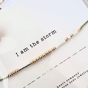 May include: A bracelet with gold and silver beads on a green string. The bracelet has the text "I am the storm" on a white card. The card also has the text "hidden heart company handcrafted morse code jewellery".