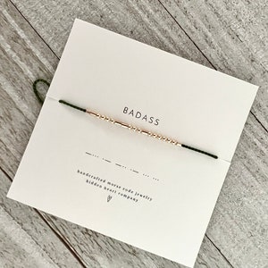 Peut inclure: Un bracelet en perles dorées avec le mot "BADASS" écrit en code Morse. Le bracelet est sur une carte blanche avec le texte "bijoux en code morse faits à la main hidden heart company" et un symbole de cœur.