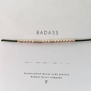 May include: A green string bracelet with gold beads spelling out the word "BADASS" in Morse code. The bracelet is handmade and comes with a card that says "handcrafted morse code jewelry hidden heart company".