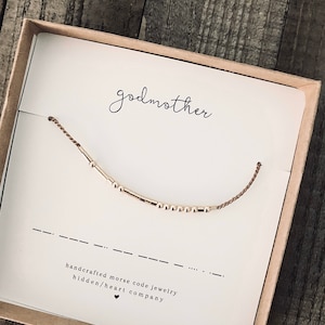 May include: A brown box with a white card inside. The card has the word "godmother" written on it in black cursive. A gold and brown bracelet with beads is displayed on the card. The card also says "handcrafted morse code jewelry hidden/heart company".