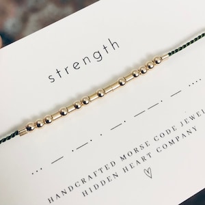 May include: A gold beaded bracelet with a green cord. The bracelet spells out the word "strength" in Morse code. The card reads "Handcrafted Morse Code Jewelry Hidden Heart Company".