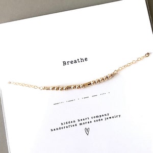 May include: Gold bracelet with a delicate chain and small beads spelling out the word "breathe" in Morse code. The bracelet is displayed on a white card with the words "hidden heart company handcrafted morse code jewelry" and a small heart.
