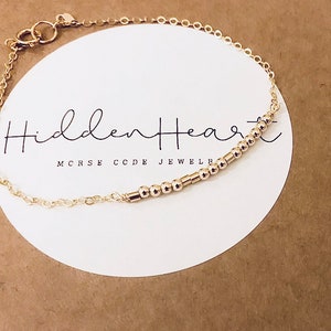 May include: A gold chain bracelet with a delicate gold bead bar charm. The charm has the text "Hidden Heart" and "Morse Code Jewellery" on a white card.