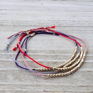 May include: A collection of colorful friendship bracelets. The bracelets feature gold beads and adjustable cords in various colors, including red, purple, pink, and blue. The bracelets are displayed on a light wood surface.
