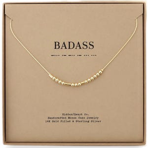 May include: A gold-colored chain necklace featuring small round and cylindrical beads arranged in a Morse code pattern. The necklace is displayed on a tan card with the text BADASS and the corresponding Morse code dots and dashes printed above.