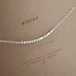 May include: A silver chain bracelet with a silver bead charm that spells out "sister" in Morse code. The bracelet is on a brown card with the text "handcrafted morse code jewelry hidden heart company".