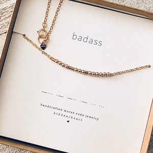 May include: Gold necklace with a small heart charm and a beaded chain that spells out "badass" in Morse code. The necklace comes in a white box with the word "badass" printed on it.