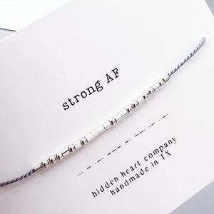 May include: A silver beaded bracelet with a gray cord and the text "strong AF" on a white card. The card also includes the text "hidden heart company handmade in TX".