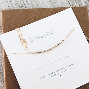 May include: Gold necklace with a delicate chain and small beads spelling out the word "breathe" in Morse code. The necklace comes in a white box with a card that reads "handcrafted morse code jewelry hidden heart".