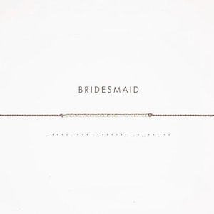 Peut inclure: Un bracelet délicat avec de petites perles claires enfilées sur un fin cordon marron. Le mot "BRIDESMAID" est imprimé au-dessus du bracelet sur fond blanc. Une ligne de points et de tirets est en dessous.