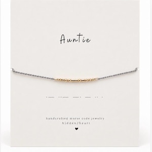 May include: A grey string bracelet with gold-colored beads arranged in Morse code, displayed on a white card featuring the text Auntie, the corresponding Morse code sequence, and the words handcrafted morse code jewelry hidden/heart.