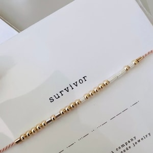 Peut inclure: Un bracelet en perles dorées et argentées avec un cordon rose. Le bracelet est accompagné d'une carte sur laquelle est écrit "survivor", ainsi que "hidden heart company handcrafted morse code jewelry".