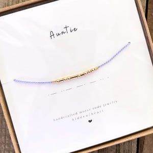 May include: A white card with the word "Auntie" written in black ink. A purple string bracelet with gold beads is displayed on the card. The bracelet is a morse code bracelet with the message "Auntie" spelled out in gold beads. The card also says "handcrafted morse code jewelry hidden/heart".