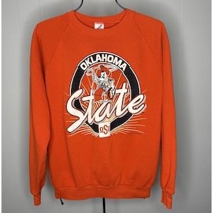 May include: Orange sweatshirt with the words "Oklahoma State" in white script and a graphic of a cowboy riding a bicycle. The sweatshirt has long sleeves and a crew neck.