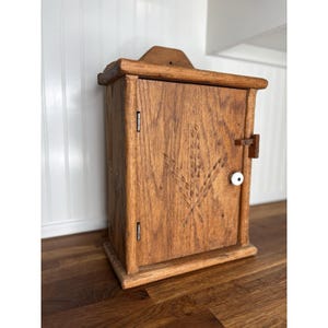 May include: A wooden bread box with a wheat stalk carving on the door. The box is made of light brown wood and has a small wooden latch and a white knob. The box is sitting on a dark brown wooden surface.