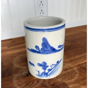 May include: A cylindrical ceramic container with a cream base and blue landscape design. The container has a blue band around the rim and a landscape scene with trees and mountains. It is likely a utensil holder or decorative piece.