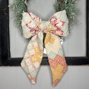 May include: A large decorative bow crafted from quilted fabric featuring a patchwork pattern of soft yellow, light blue, and muted pink floral squares. The bow has two long, trailing tails and is positioned over a green leafy wreath.