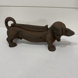 May include: A dark brown, cast iron dachshund-shaped boot scraper or doorstop. The object features a long, low-slung body, a curled tail, and a textured, weathered metal surface. The design includes a central recessed area along the back.