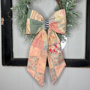 May include: A large decorative bow made from quilted fabric featuring a patchwork of various patterns including stripes, floral prints, and solid muted tones in shades of pink, green, red, and blue, centered on a green leafy wreath.