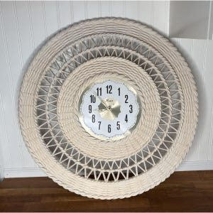 May include: A round, cream-colored woven wall clock with a gold-toned clock face. The clock features a woven design with concentric circles and geometric patterns. The clock face displays black numerals and hands, with the brand name "Xemi".