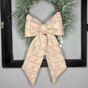 May include: A large decorative bow crafted from quilted fabric featuring a cream and faded red geometric triangle pattern. The bow has two long, pointed tails and is positioned over a circular wreath made of green leaves and thin brown twigs.