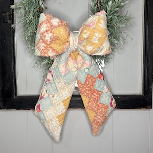 May include: A large decorative bow crafted from quilted fabric featuring a patchwork pattern in soft shades of yellow, peach, light blue, and white. The bow is positioned over a wreath made of green foliage against a black window frame.