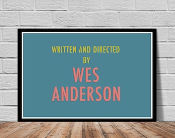 May include: Framed poster with the text "WRITTEN AND DIRECTED BY WES ANDERSON" in yellow and coral-pink against a teal background. The poster is in a black frame and is displayed on a wooden floor against a white brick wall.