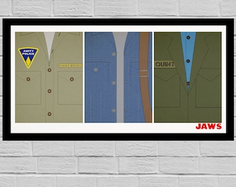 May include: A framed print featuring three panels of clothing from the movie "Jaws." The panels show the uniforms of Chief Brody, Quint, and a denim jacket. The Amity Police badge and the word "JAWS" are visible.